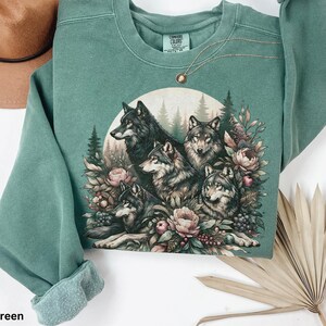 May include: Light green sweatshirt with a graphic print of a pack of wolves surrounded by flowers and foliage.