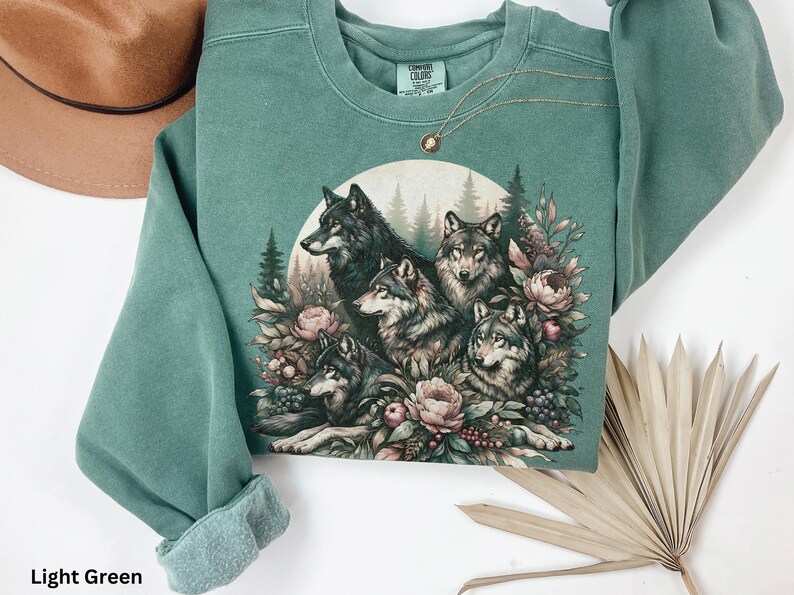 May include: Light green sweatshirt with a graphic print of a pack of wolves surrounded by flowers and foliage.