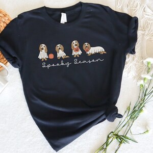 May include: Black t-shirt featuring a Halloween-themed design. The design includes illustrations of dogs dressed as ghosts and holding pumpkins. The phrase "Spooky Season" is written in a script font below the dogs. The shirt is a dark color.