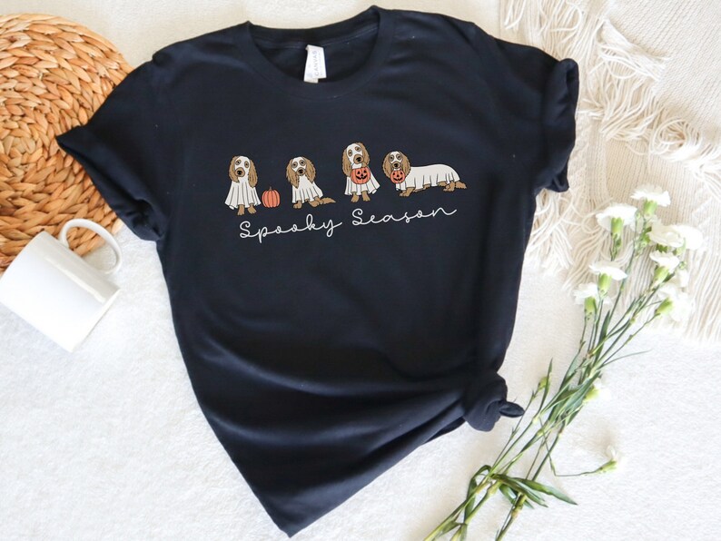 May include: Black t-shirt featuring a Halloween-themed design. The design includes illustrations of dogs dressed as ghosts and holding pumpkins. The phrase "Spooky Season" is written in a script font below the dogs. The shirt is a dark color.