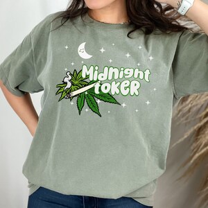 May include: Sage green t-shirt with the text "Midnight Toker" in green and white, featuring a cartoon moon, stars, and cannabis leaf design. The shirt is a relaxed fit.