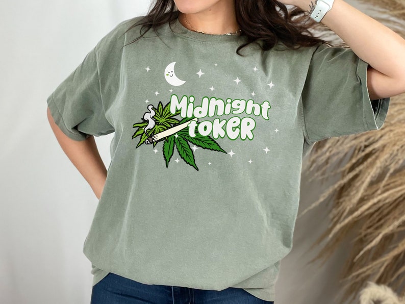 May include: Sage green t-shirt with the text "Midnight Toker" in green and white, featuring a cartoon moon, stars, and cannabis leaf design. The shirt is a relaxed fit.