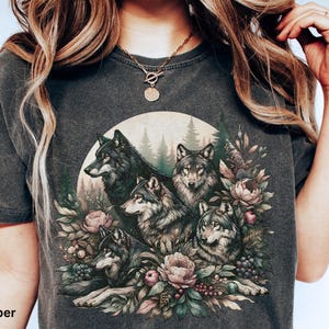 May include: A dark gray t-shirt with a graphic print of five wolves surrounded by flowers and greenery. The wolves are depicted in a realistic style with detailed fur and expressions.