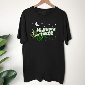 May include: Black t-shirt with a graphic design featuring a marijuana leaf, a lit joint, a crescent moon, stars, and the text "Midnight Toker" in green and white. The shirt is hanging on a wooden hanger.