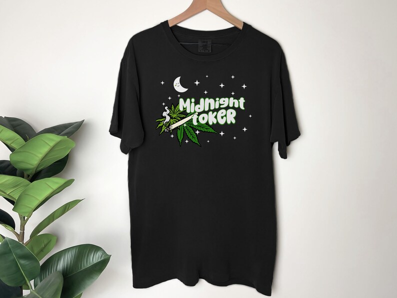 May include: Black t-shirt with a graphic design featuring a marijuana leaf, a lit joint, a crescent moon, stars, and the text "Midnight Toker" in green and white. The shirt is hanging on a wooden hanger.