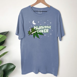 May include: A blue t-shirt with the words "Midnight Toker" in green and white, along with a cannabis leaf and a lit joint. The design includes a crescent moon and stars.
