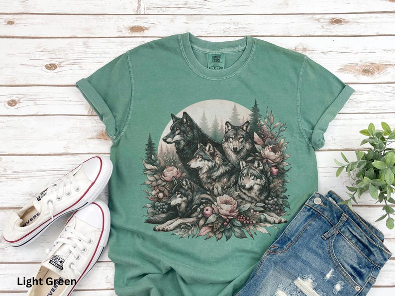 May include: A light green t-shirt with a graphic print of a group of wolves surrounded by flowers and foliage. The wolves are in shades of gray and brown, and the flowers are in shades of pink and purple.