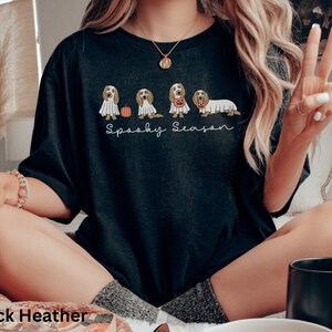 May include: Black heather t-shirt with a "Spooky Season" design. The design features a line of dogs dressed in Halloween costumes, including a ghost and a pumpkin. The shirt is a comfortable, casual style.