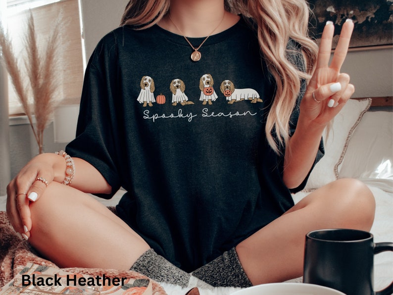 May include: Black heather t-shirt with a "Spooky Season" design. The design features a line of dogs dressed in Halloween costumes, including a ghost and a pumpkin. The shirt is a comfortable, casual style.