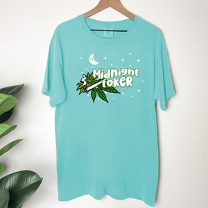 May include: A turquoise t-shirt with the words "Midnight Toker" in green and white, along with a marijuana leaf and a lit joint graphic. The design includes a crescent moon and stars.