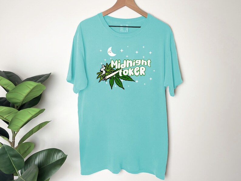 May include: A turquoise t-shirt with the words "Midnight Toker" in green and white, along with a marijuana leaf and a lit joint graphic. The design includes a crescent moon and stars.