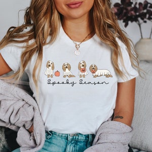 May include: White t-shirt featuring a Halloween-themed design. The graphic includes several dogs dressed in costumes, such as ghosts and holding jack-o'-lanterns, with the words "Spooky Season" written below. The shirt is a casual, festive apparel item.
