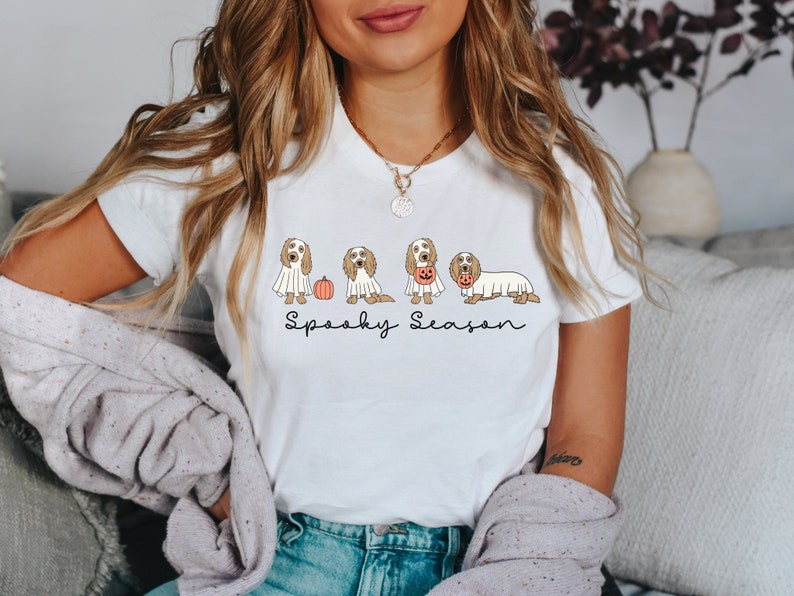 May include: White t-shirt featuring a Halloween-themed design. The graphic includes several dogs dressed in costumes, such as ghosts and holding jack-o'-lanterns, with the words "Spooky Season" written below. The shirt is a casual, festive apparel item.