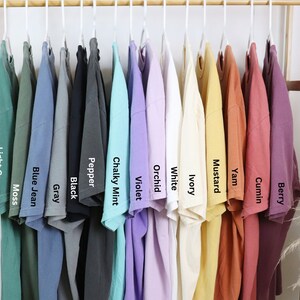 May include: A rack of 12 t-shirts in various colors, including light green, blue jean, gray, black, pepper, chalky mint, violet, orchid, white, ivory, mustard, yam, cumin, and berry.