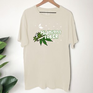 May include: Cream-colored t-shirt with the words "Midnight Toker" in green, accompanied by a marijuana leaf and a lit joint graphic. A crescent moon and stars are above the text. The shirt is hanging on a wooden hanger.