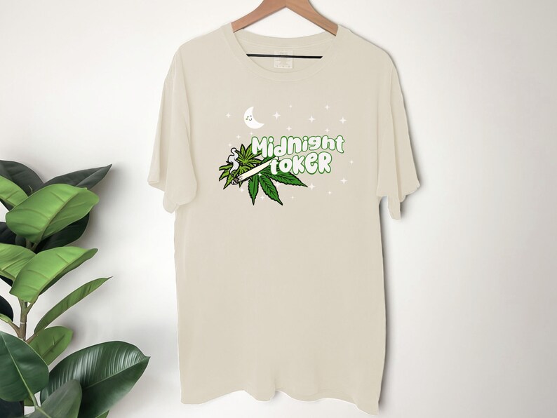 May include: Cream-colored t-shirt with the words "Midnight Toker" in green, accompanied by a marijuana leaf and a lit joint graphic. A crescent moon and stars are above the text. The shirt is hanging on a wooden hanger.