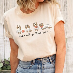 May include: Cream-colored t-shirt featuring a Halloween-themed graphic. The design includes illustrations of dogs dressed as ghosts and holding pumpkins, with the words "Spooky Season" in a handwritten font.