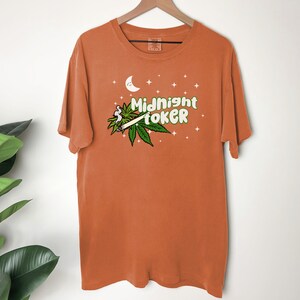 May include: Orange t-shirt with the text "Midnight Toker" in green and white, featuring a cartoon joint and cannabis leaves. The design includes a crescent moon and stars. The shirt is hanging on a wooden hanger.