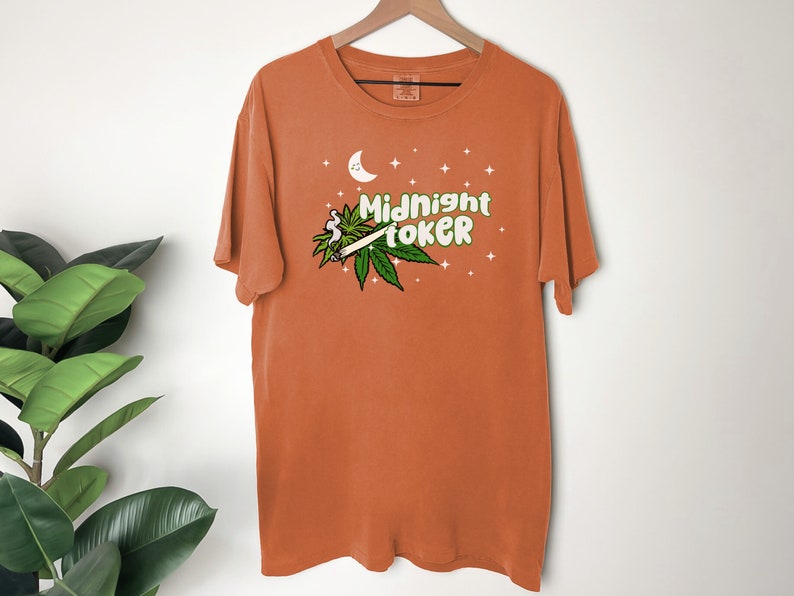 May include: Orange t-shirt with the text "Midnight Toker" in green and white, featuring a cartoon joint and cannabis leaves. The design includes a crescent moon and stars. The shirt is hanging on a wooden hanger.