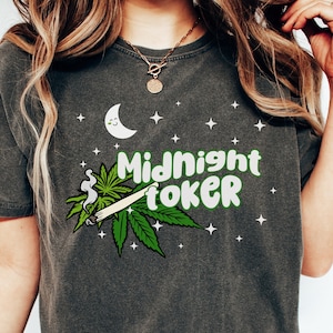 May include: Dark grey t-shirt with a graphic design featuring a crescent moon, stars, and cannabis leaves. The words "Midnight Toker" are written in green and white. The shirt has a vintage, worn look.