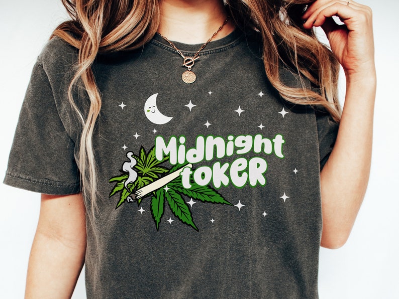 May include: Dark grey t-shirt with a graphic design featuring a crescent moon, stars, and cannabis leaves. The words "Midnight Toker" are written in green and white. The shirt has a vintage, worn look.