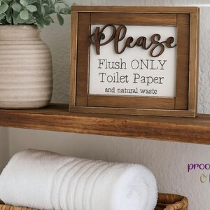 May include: A wooden framed sign with the words "Please Flush ONLY Toilet Paper and natural waste" in black and brown lettering. The sign is on a wooden shelf with a rolled white towel and a decorative vase.