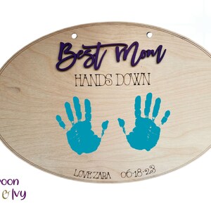 May include: A wooden oval sign with the text "Best Mom Hands Down" in black lettering. Two teal handprints are on the sign with the date "06-18-23" and the text "Love Zara" below the handprints.