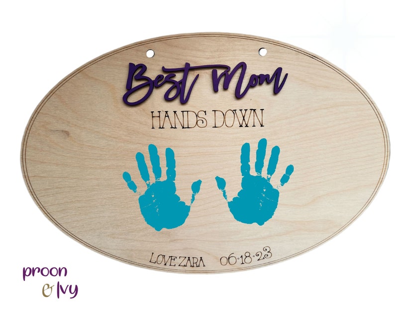 May include: A wooden oval sign with the text "Best Mom Hands Down" in black lettering. Two teal handprints are on the sign with the date "06-18-23" and the text "Love Zara" below the handprints.