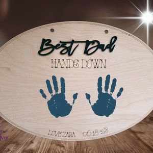 May include: Oval wooden sign with the text "Best Dad Hands Down" in black lettering. Two blue handprints are on the sign with the text "Love Zara 06-18-23" below.