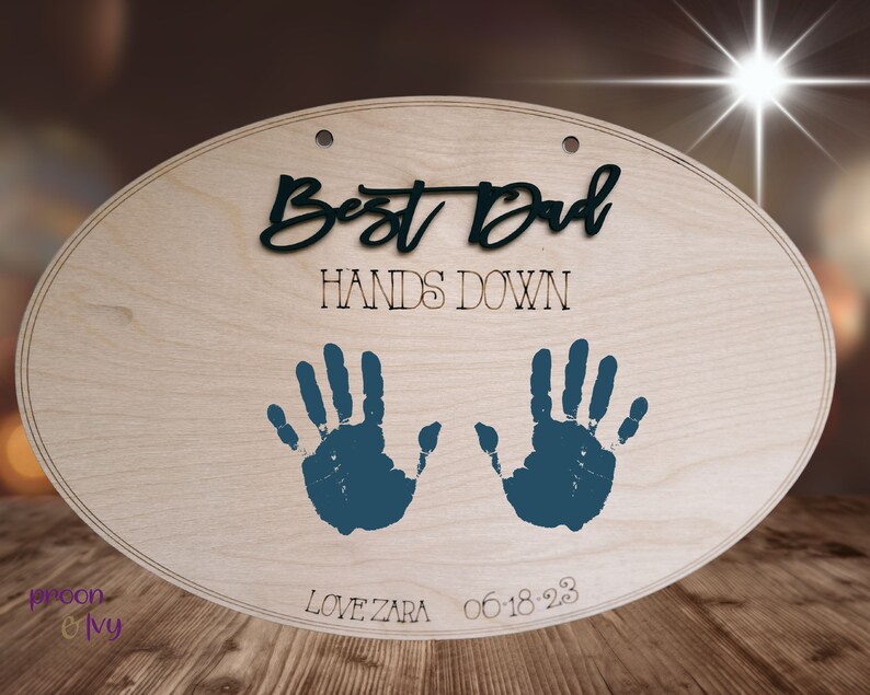 May include: Oval wooden sign with the text "Best Dad Hands Down" in black lettering. Two blue handprints are on the sign with the text "Love Zara 06-18-23" below.