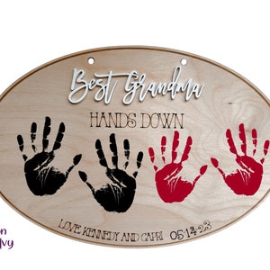 May include: Wooden oval sign with the text "Best Grandma Hands Down" in white and black lettering. Three handprints are visible, two black and one red. The text "Love Kennedy and Capri 05.14.23" is also visible.