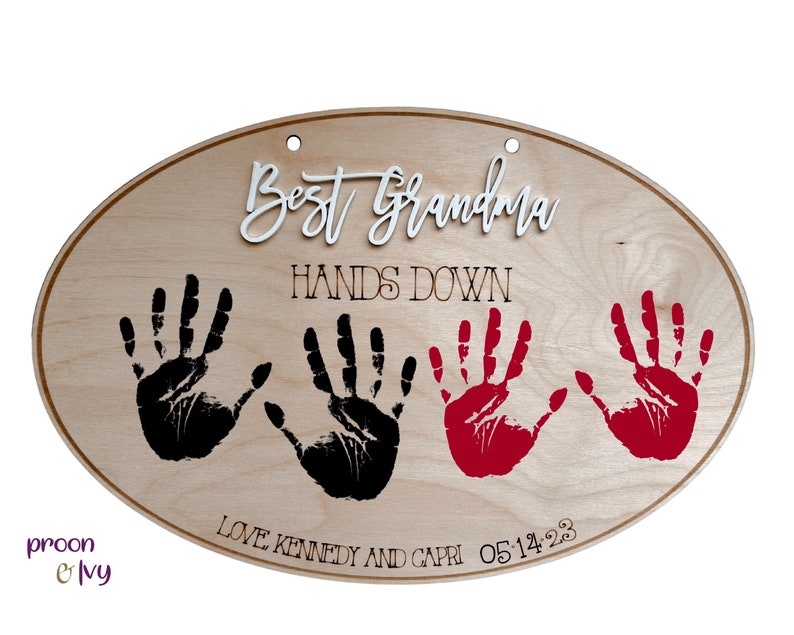 May include: Wooden oval sign with the text "Best Grandma Hands Down" in white and black lettering. Three handprints are visible, two black and one red. The text "Love Kennedy and Capri 05.14.23" is also visible.