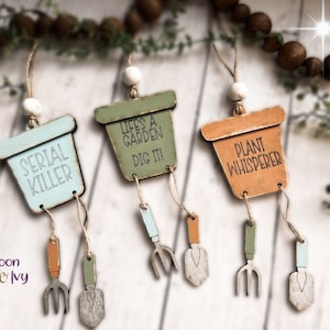 May include: Three wooden pot-shaped ornaments with hanging gardening tools. The pots are painted in different colors and have the following text: "Serial Killer", "Life's a Garden Dig It!" and "Plant Whisperer".