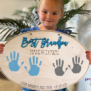 May include: A wooden oval sign with the text "Best Grandpa Hands Down" in blue and black lettering. Two sets of blue and gray handprints are on the sign with the text "Love Kennedy and Capri 06.18.23" below the handprints.