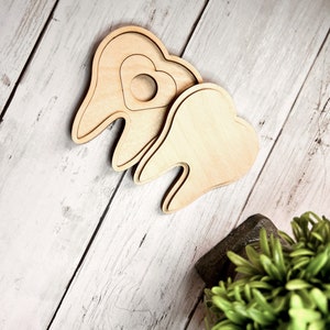 May include: Two wooden teeth shapes, one with a heart cutout, on a white wood background.
