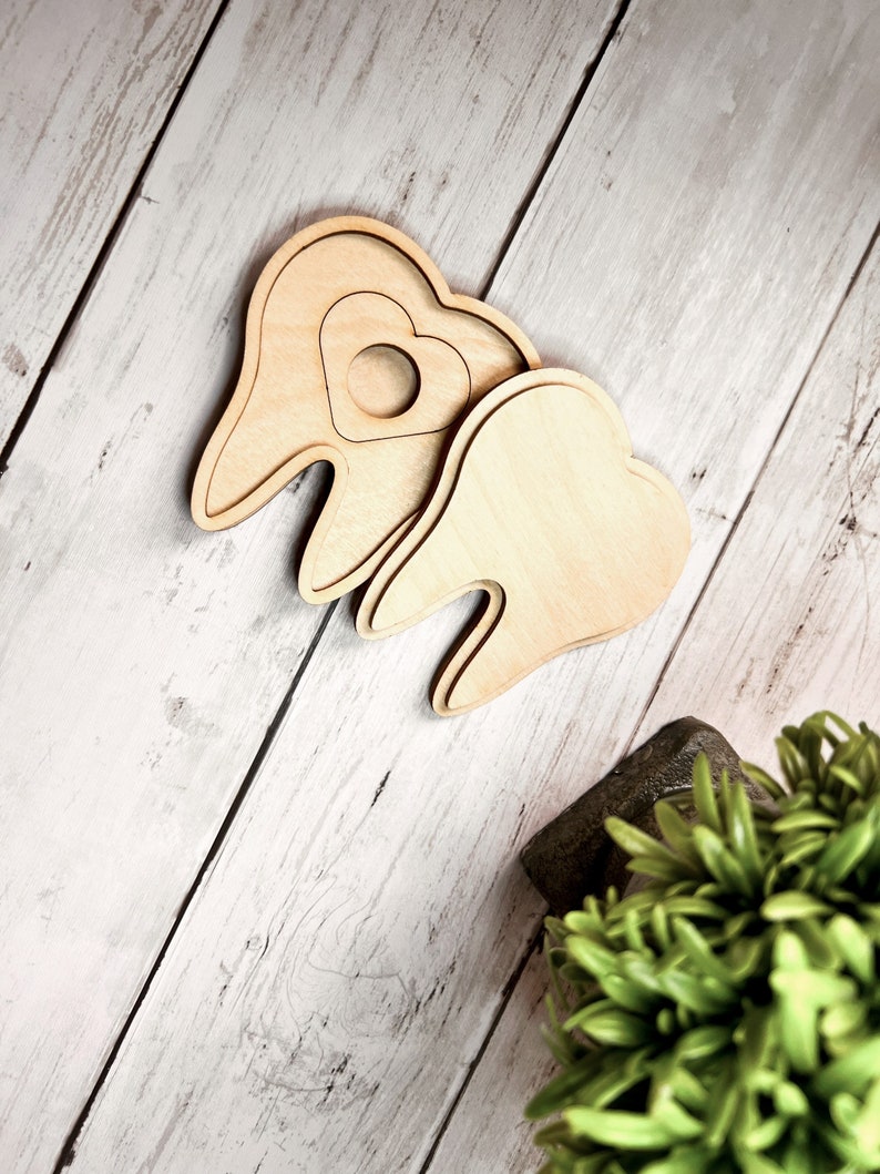 May include: Two wooden teeth shapes, one with a heart cutout, on a white wood background.