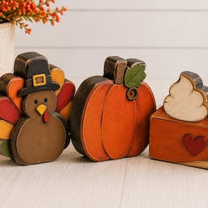 May include: Three wooden tabletop decorations featuring a brown turkey wearing a black hat, a round orange pumpkin with a green leaf, and a slice of orange pumpkin pie topped with white whipped cream and a small red heart accent.