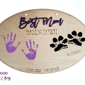 May include: Wooden oval sign with the text "Best Mom Hands Down" in purple and black lettering. The sign also features two purple handprints, two black paw prints, and the text "& Dewey! Love Zara 06.18.23"