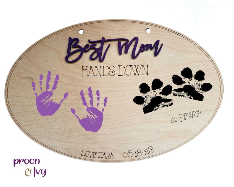 May include: Wooden oval sign with the text "Best Mom Hands Down" in purple and black lettering. The sign also features two purple handprints, two black paw prints, and the text "& Dewey! Love Zara 06.18.23"