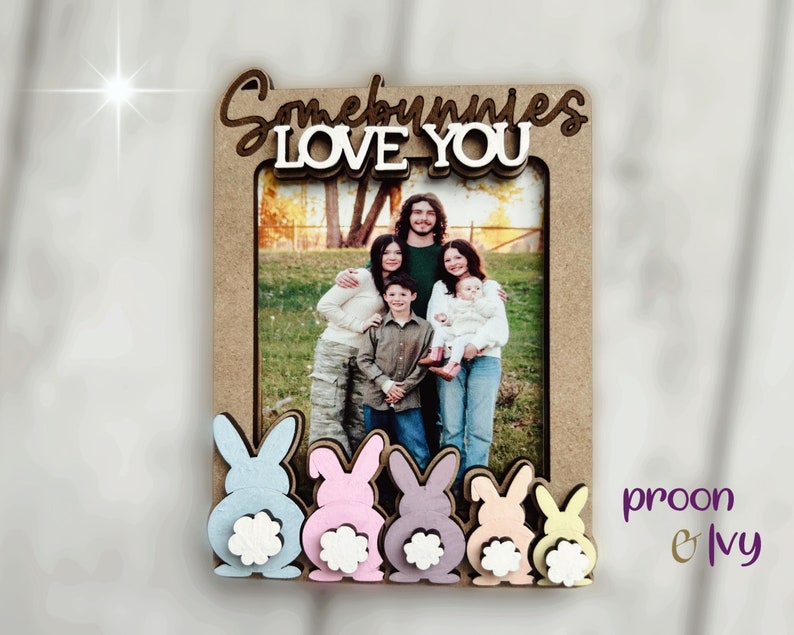 Peut inclure: Un cadre photo en bois avec l'inscription "Somebunnies LOVE YOU" en &eacute;criture cursive. Le cadre a une ouverture rectangulaire pour une photo et est d&eacute;cor&eacute; de d&eacute;coupes de lapins aux couleurs pastel en bas. Le cadre est de couleur marron clair.