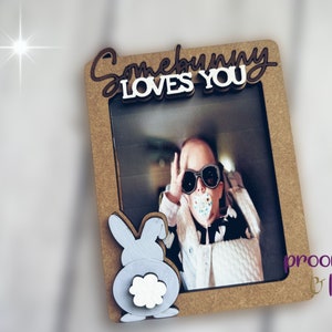 Peut inclure: Un cadre photo rectangulaire avec les mots "Somebunny Loves You" en marron et blanc. Le cadre a une d&eacute;coupe pour une photo et un motif de lapin d&eacute;coratif. La photo montre une personne portant des lunettes de soleil.