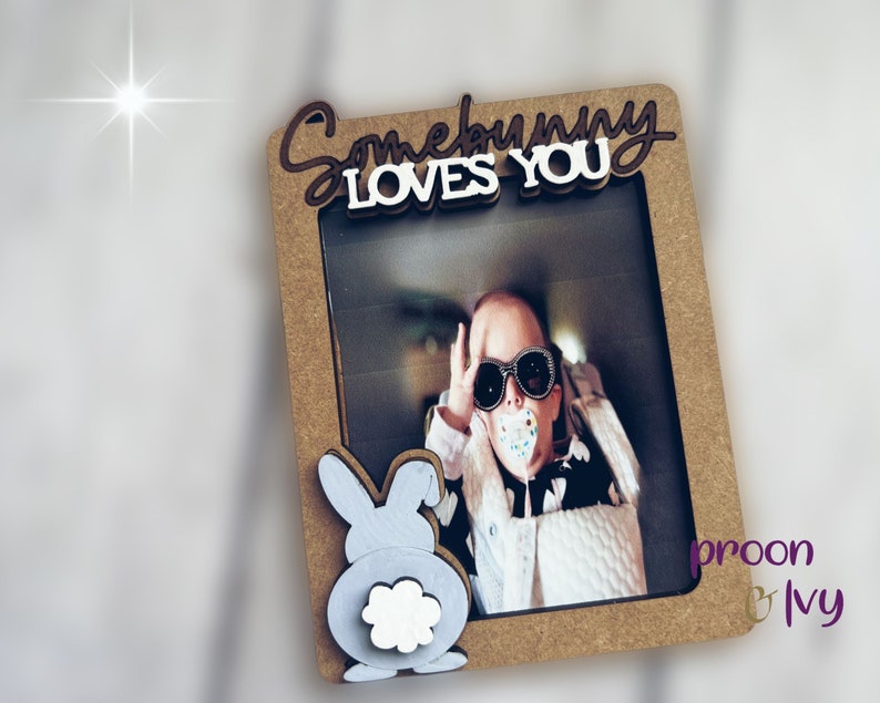 Peut inclure: Un cadre photo rectangulaire avec les mots "Somebunny Loves You" en marron et blanc. Le cadre a une d&eacute;coupe pour une photo et un motif de lapin d&eacute;coratif. La photo montre une personne portant des lunettes de soleil.