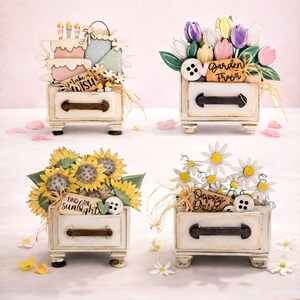 May include: Four decorative wooden drawer boxes, each with a different floral or celebratory theme. One features a cake and gifts, another tulips, a third sunflowers, and the last daisies. Each box has a decorative handle and is painted white.