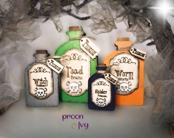 May include: Four small glass bottles with cork stoppers and labels. The bottles are painted in different colors: gray, green, blue, and orange. The labels are made of wood and have a skull and crossbones design. The labels read "Witch Potion", "Toad Brains", "Spider Venom", and "Worm Worts".