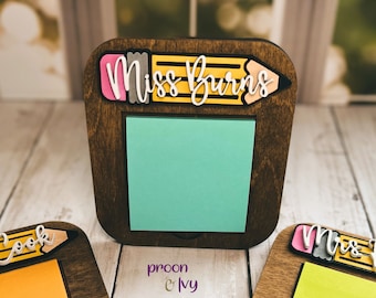 May include: A wooden pencil-shaped note holder with a light blue sticky note. The pencil has a pink eraser and the text "Miss Burns" is written on the pencil.