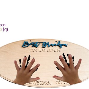 May include: A wooden oval sign with the text "Best Grandpa Hands Down" in blue lettering. Two black handprints are on the sign with the text "Love Zara 06.10" below the handprints.