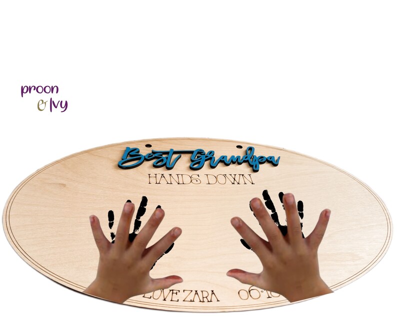 May include: A wooden oval sign with the text "Best Grandpa Hands Down" in blue lettering. Two black handprints are on the sign with the text "Love Zara 06.10" below the handprints.