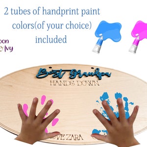 May include: Wooden oval sign with the text "Best Grandpa Hands Down" and space for two handprints. The sign is light brown and has a natural wood grain. Two tubes of paint are included, one blue and one pink.