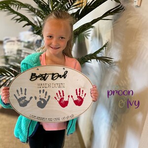 May include: A wooden oval sign with the text "Best Dad Hands Down" and three handprints in gray and red. The sign also includes the text "Love Kennedy and Gwen 06-18-23".