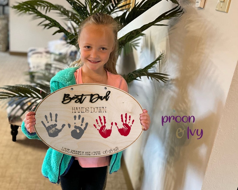 May include: A wooden oval sign with the text "Best Dad Hands Down" and three handprints in gray and red. The sign also includes the text "Love Kennedy and Gwen 06-18-23".